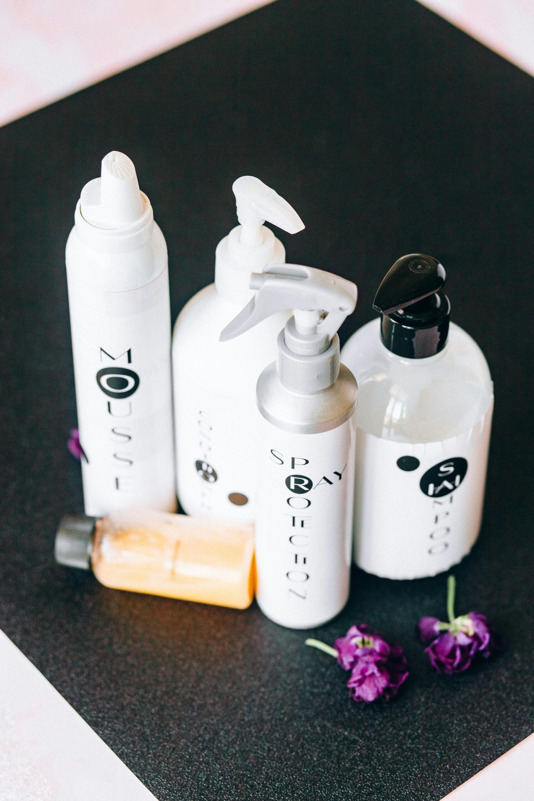 Elegant arrangement of hair care product bottles with vibrant purple flowers on a dark background.